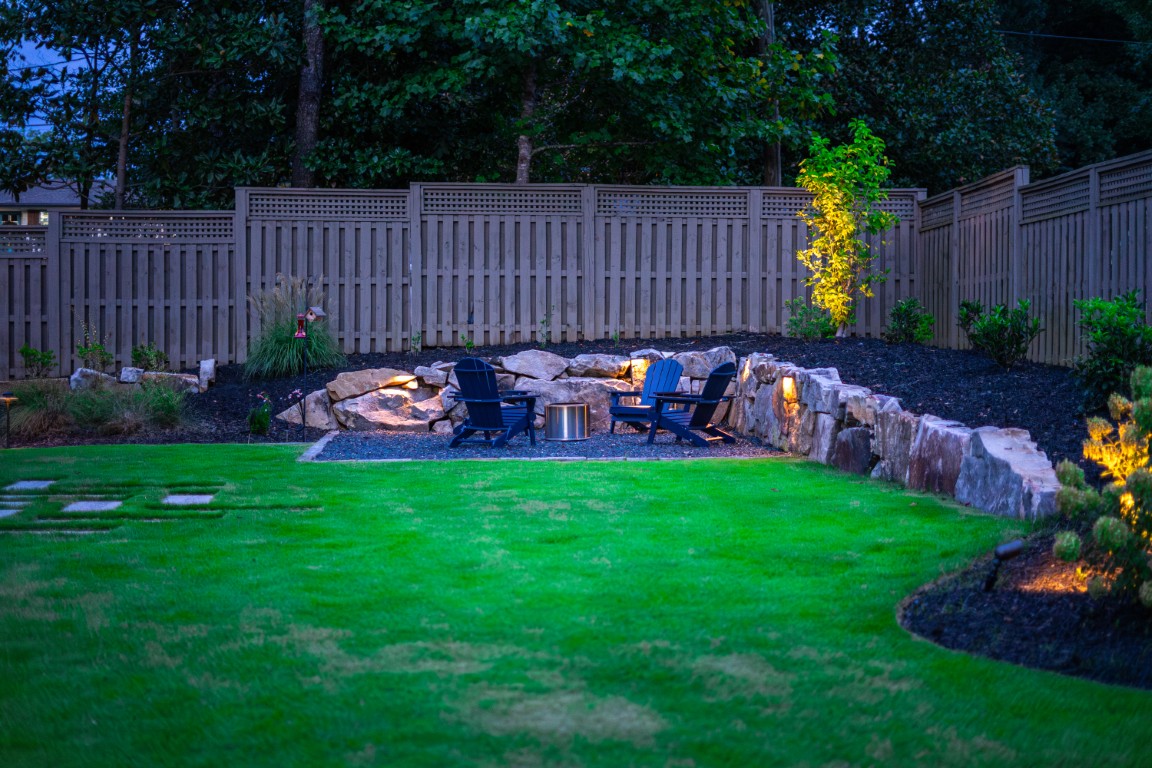 At dusk, a backyard features green grass, a wooden fence, stone landscaping, four Adirondack chairs around a fire pit, and soft outdoor lights.
