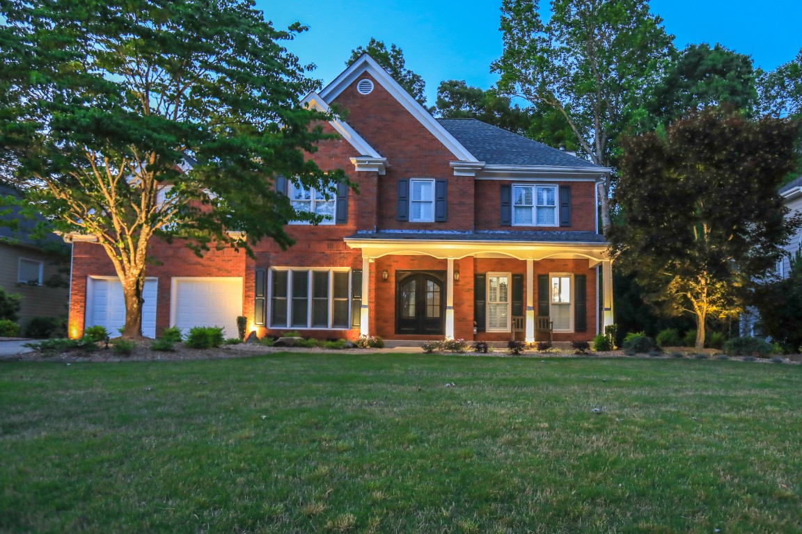 A two-story brick house with a front porch, double doors, and two-car garage is lit by exterior lights at dusk and surrounded by trees.
