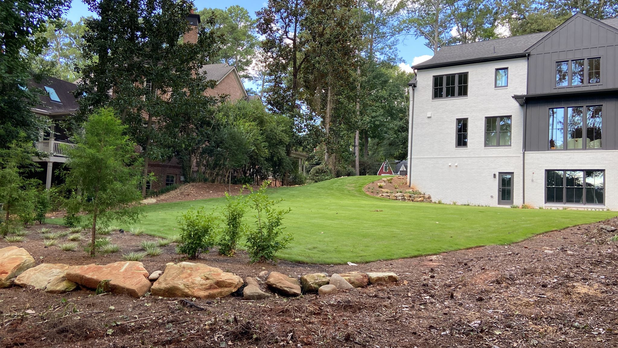 A modern white house with large windows faces a green lawn bordered by rocks and shrubs, with mature trees and another house in the background.