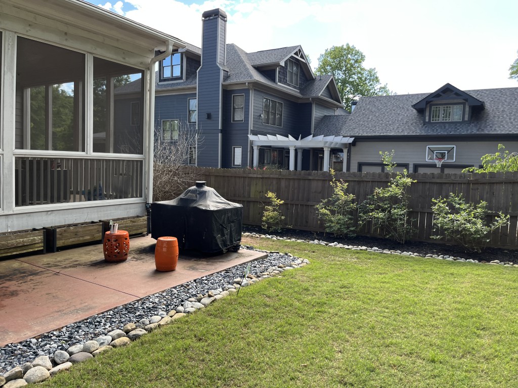 A backyard features a covered grill, two orange stools, a lawn with shrubs, a wooden fence, two gray houses, and a basketball hoop in back.