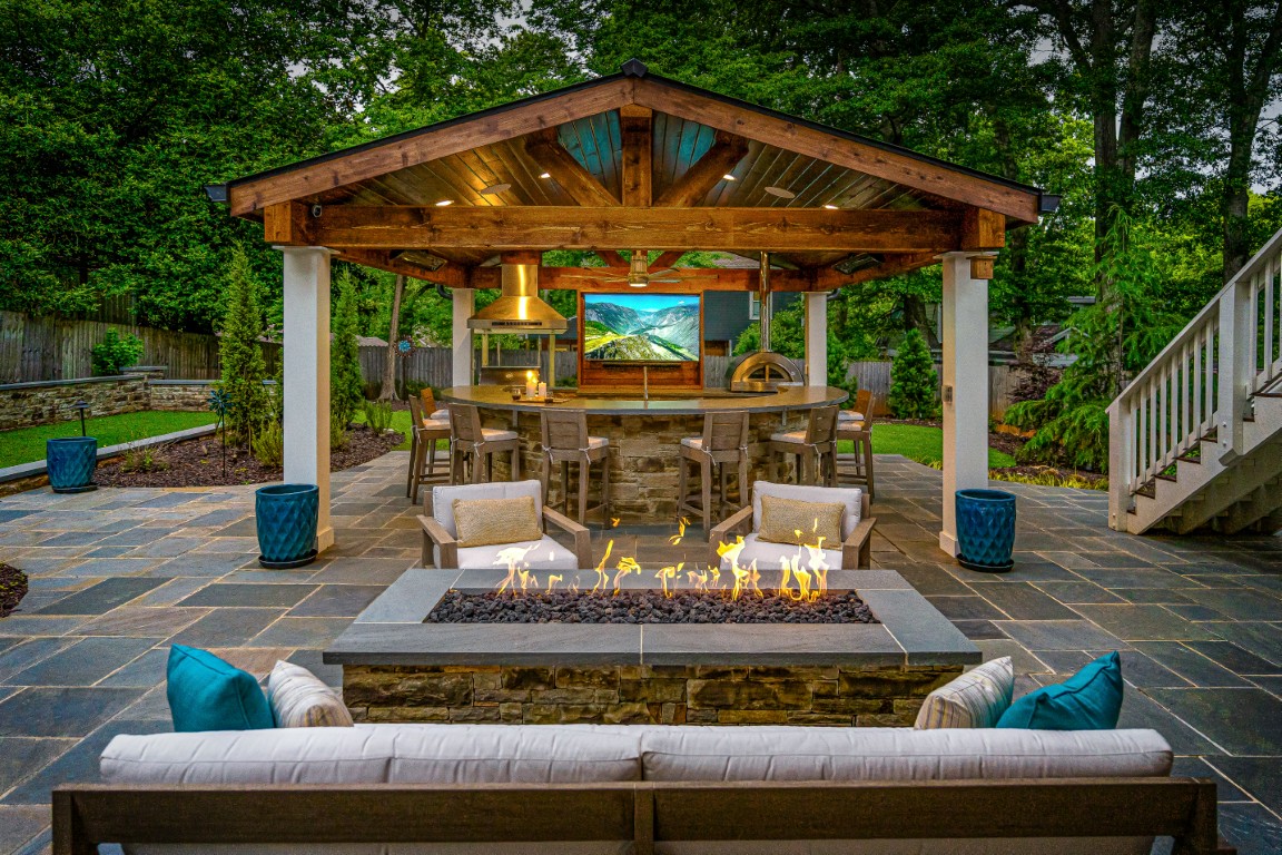 A cozy outdoor patio features a fire pit, sofa, covered bar with stools, hanging lights, wooden beams, TV, greenery, and stone paving.