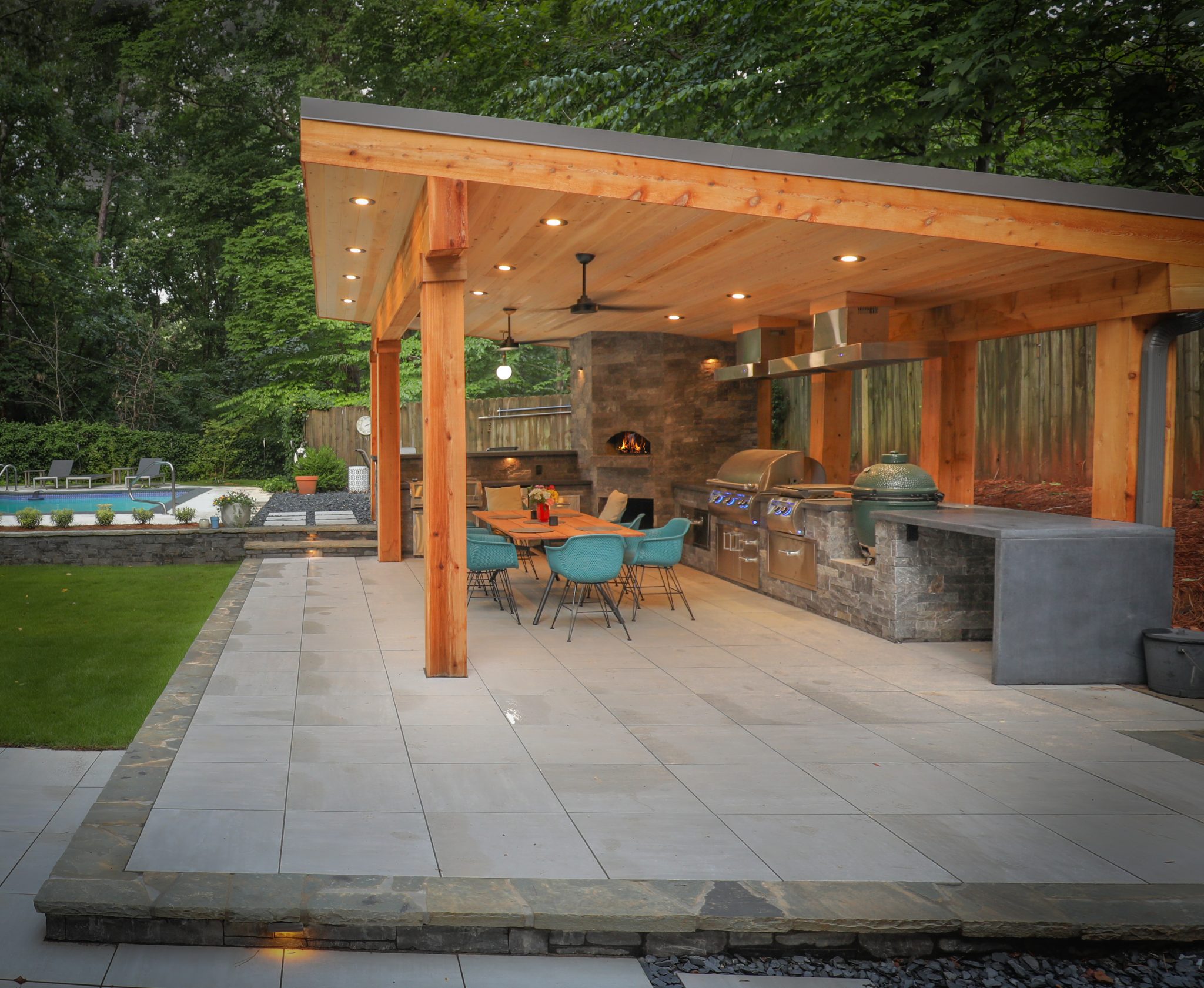 Modern outdoor kitchen under a wooden pavilion with grill, pizza oven, dining table, turquoise chairs, and ceiling fan by a pool and greenery.