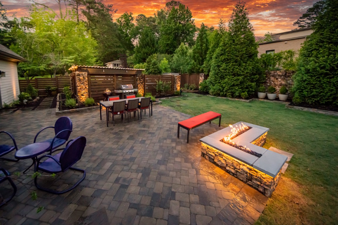 Backyard patio with a dining table, grill, blue chairs, and a red bench by a rectangular fire pit, surrounded by trees at sunset.