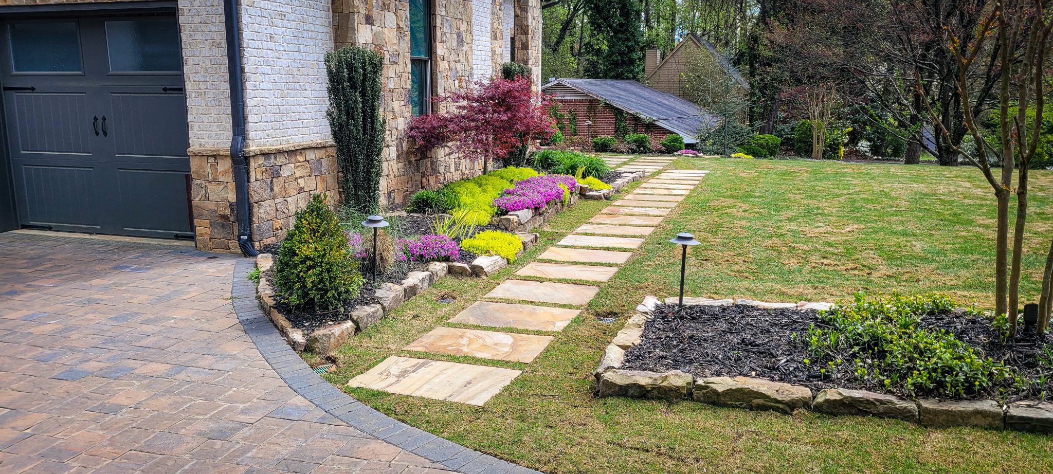 A stone walkway leads past a garden bed of purple and yellow flowers along the side of a stone house.