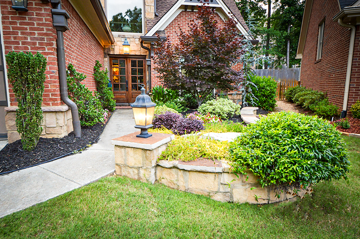 A red brick house entrance in Atlanta features a stone pathway, a low stone planter, and various shrubs and trees in the front garden.