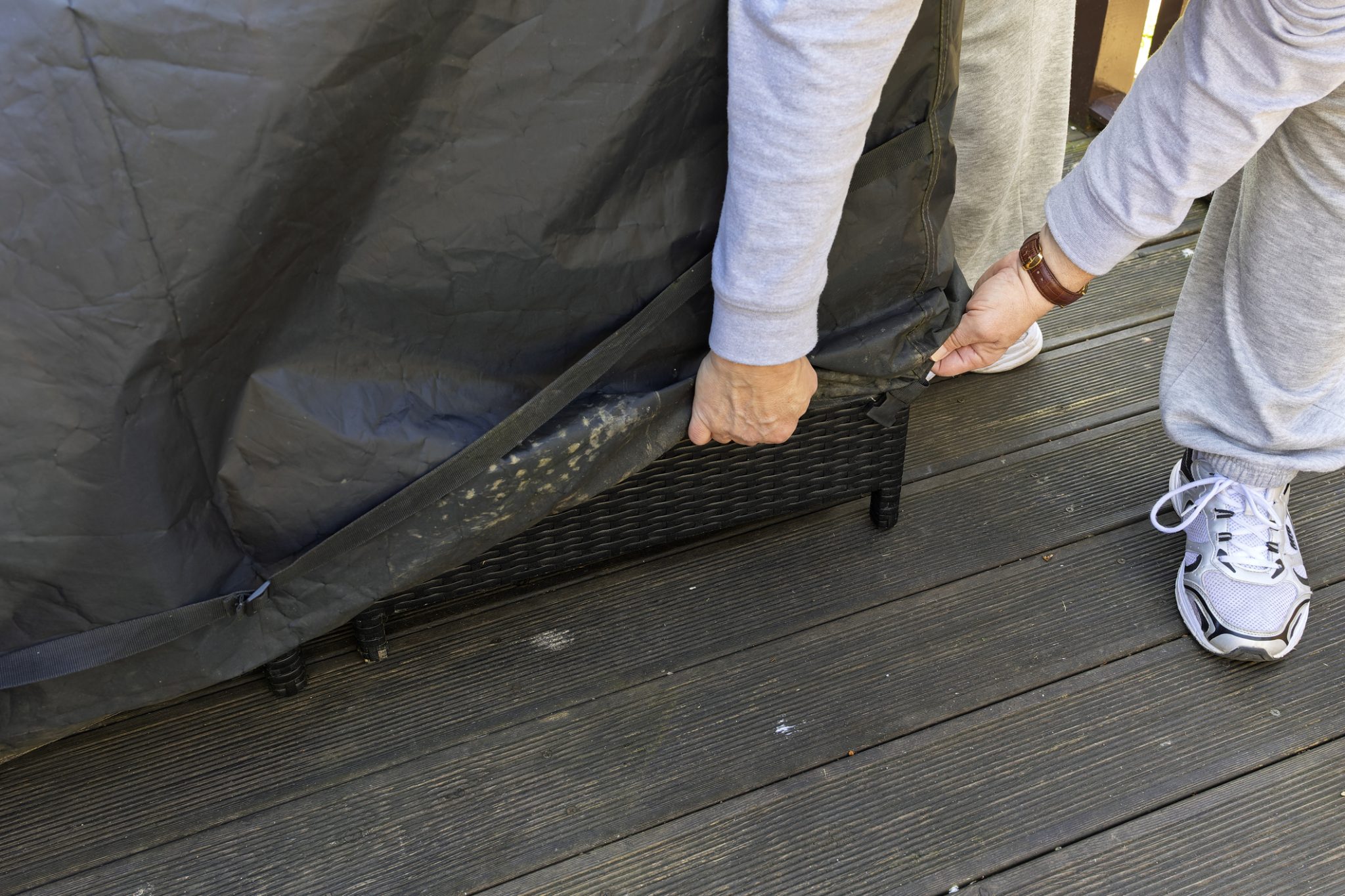 A person pulls a black cover off a wicker-style patio chair on a wooden deck.