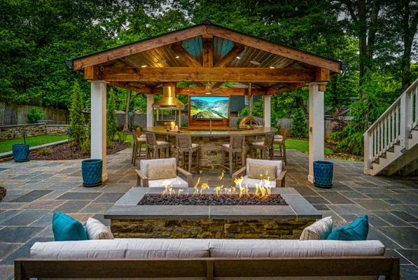 An outdoor patio features a fire pit facing a covered kitchen area complete with a bar, stools, and a television.